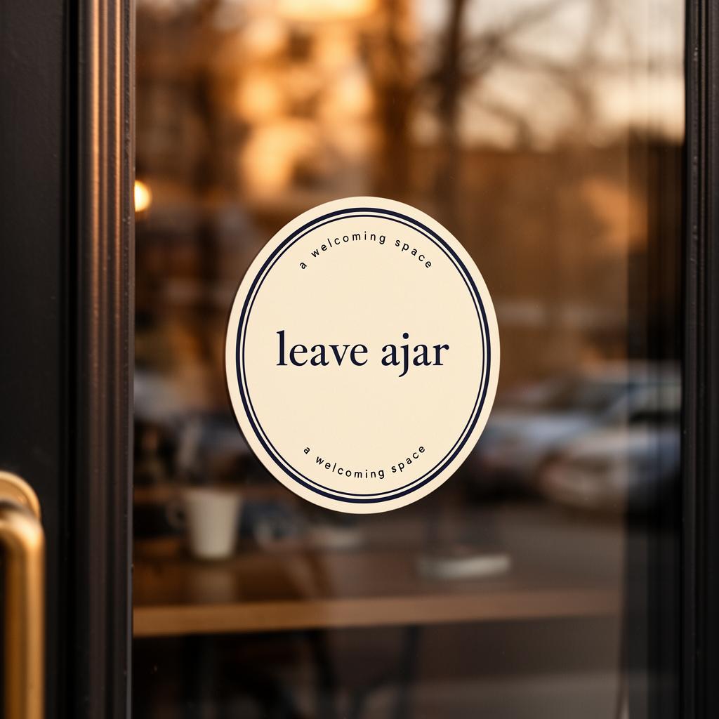 A circular cream window sticker reading 'leave ajar — a welcoming space' on the glass door of a café.